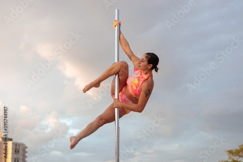 Young Middle Eastern pole dancer, holding a pose on a pole set outdoors. Tel Aviv buildings in the far background.