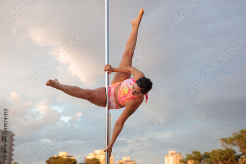 Young Middle Eastern pole dancer, holding a pose on a pole set outdoors. Tel Aviv buildings in the far background.