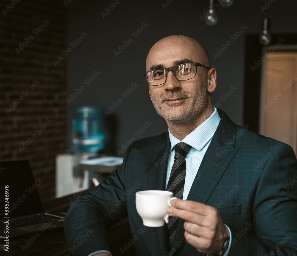Mature business man in a business suit, holding a cup of coffee in a office building cafeteria having and looking at camera wearing expensive suit and eye glasses. Business concept. Mature business man in a business suit, holding a cup of coffee in a office building cafeteria having and looking at camera wearing expensive suit and eye glasses. Business concept.