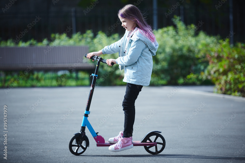 Happy beautiful girl is riding along the street on the kick scooter on a windy day. Baby having free time playing. The concept of a healthy lifestyle. Kids sport Happy beautiful girl is riding along the street on the kick scooter on a windy day. Baby having free time playing. The concept of a healthy lifestyle. Kids sport