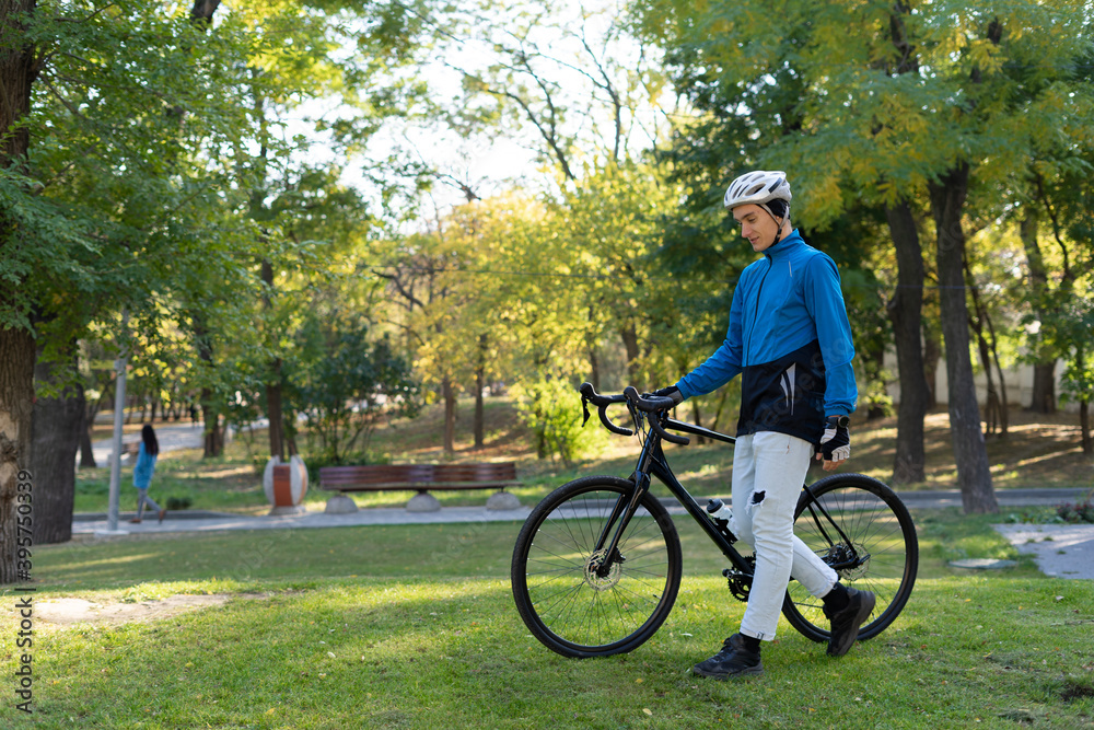 a young man of European appearance walks through the park and rolls his bike alongside.