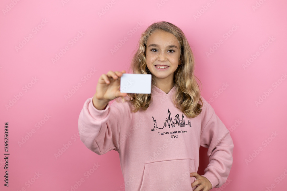 Young beautiful child girl standing over isolated pink background smiling and holding white card Young beautiful child girl standing over isolated pink background smiling and holding white card