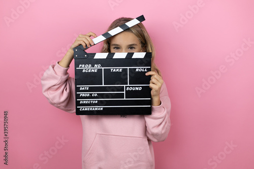Young beautiful child girl standing over isolated pink background holding clapperboard very happy
