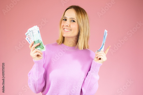 Portrait of a cheerful young woman holding money banknotes and celebrating isolated over pink background