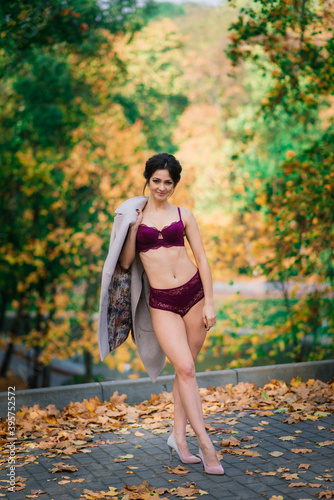 Fototapeta Naklejka Na Ścianę i Meble -  Young beautiful dark-haired woman with slender figure in lingerie and classic coat in autumn park