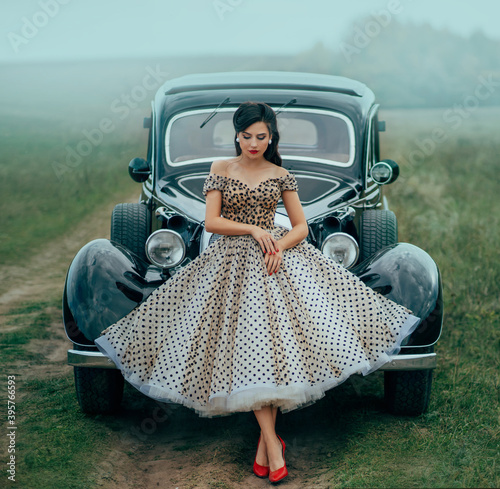 Young beautiful sexy woman in pin-up style clothes posing near black retro car. Polka dot white dress, vintage hairstyle, red high heels. Background road green nature fog. Girl fashion model driver photo