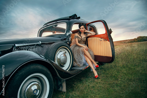 Εκτύπωση καμβά Young beautiful sexy woman in pin-up style clothes sits posing in black retro car