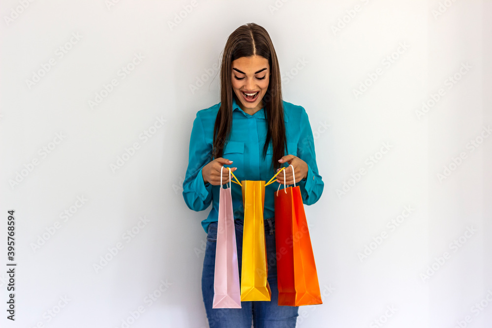 Caucasian woman holding colorful shopping bags, posing on white background. Girl with big smile enjoying shopping sales. Shocked young woman in trendy outfit looking at purchases inside paper bag.