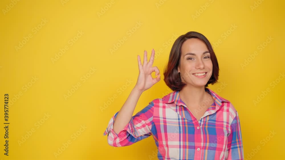 OK sign with one raised hand smile beautiful girl dressed in a plaid shirt and dark hair on a yellow background. Human emotions, facial expression concept. High quality 4k footage. OK sign with one raised hand smile beautiful girl dressed in a plaid shirt and dark hair on a yellow background. Human emotions, facial expression concept. High quality 4k footage.