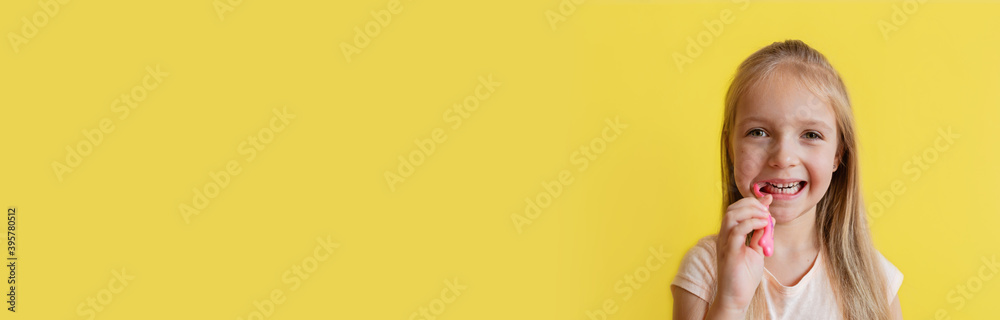 Cute little caucasian girl with blonde hair cleaning teeth with toothbrush on yellow background. Horizontal banner for web design. Studio shot with copy space. Dental hygiene and healthy concept
