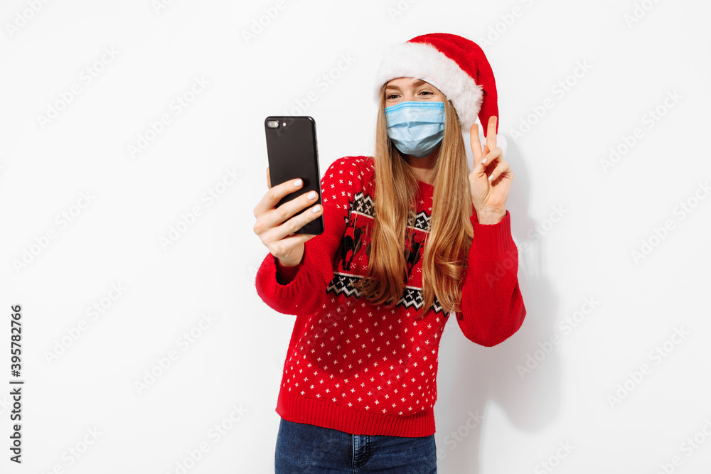 Young happy woman in a red Santa Claus hat and a medical mask on her face, waving, making a video call on a smartphone on Christmas eve, on a white background Young happy woman in a red Santa Claus hat and a medical mask on her face, waving, making a video call on a smartphone on Christmas eve, on a white background
