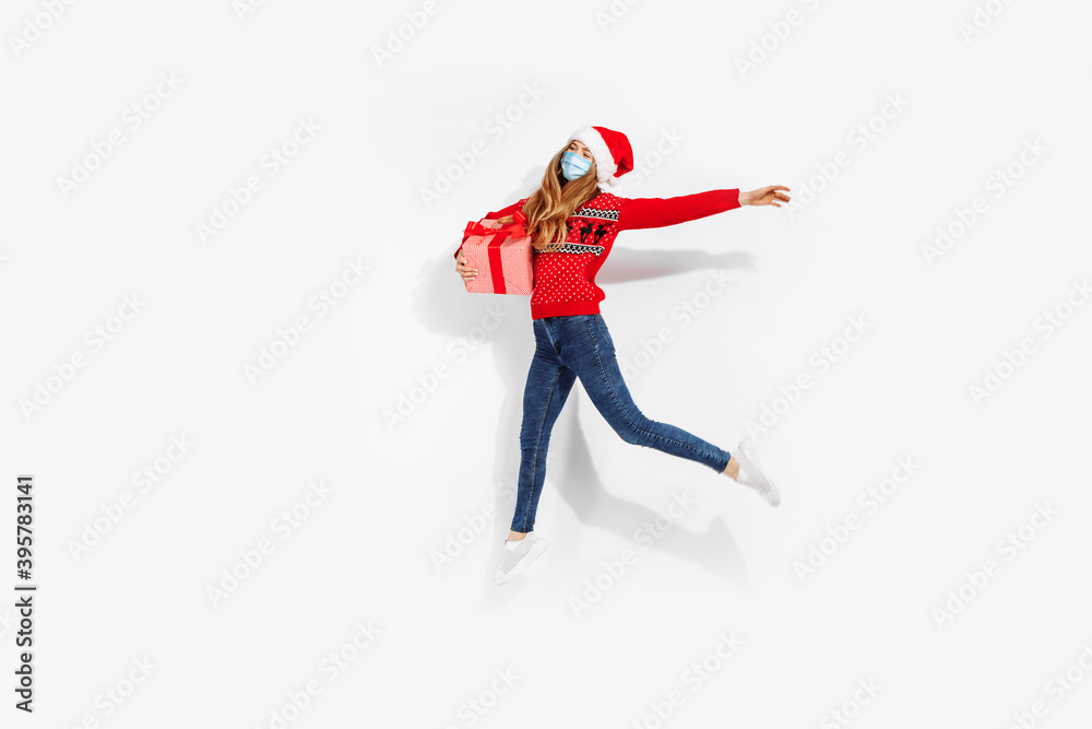 Fototapeta premium Full length, attractive young woman in christmas sweater, santa claus hat and medical face mask holding gift box, jumping cheerfully against white background