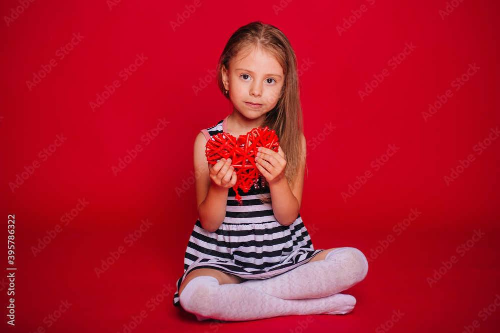 A little girl with blond hair in a striped dress holds a red heart in her hands. Child on a red background. A little girl with blond hair in a striped dress holds a red heart in her hands. Child on a red background.