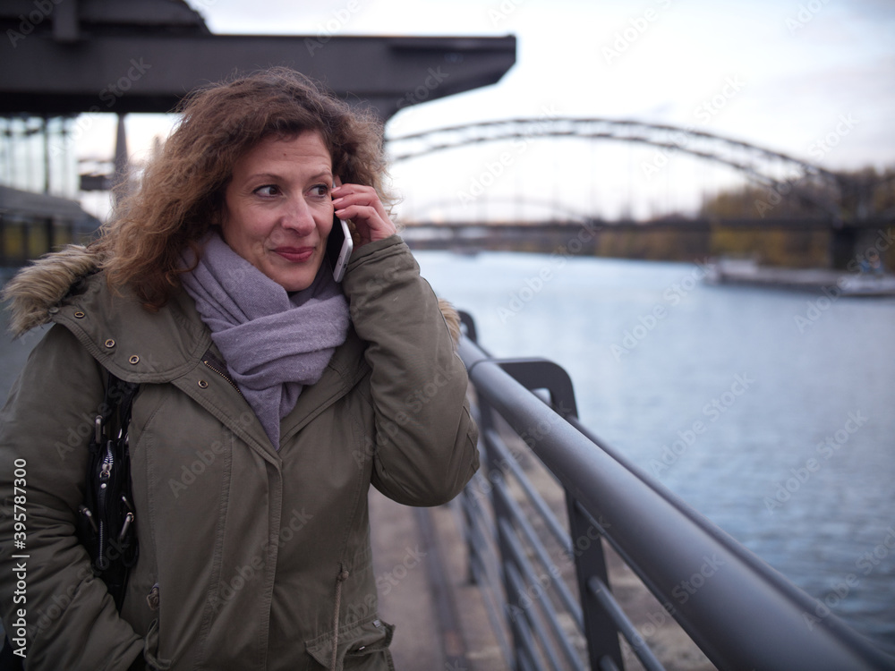 Fototapeta premium Woman stands next to a the river Main and is on the phone. She smiles and seems happy. City Frankfurt am Main during autumn.