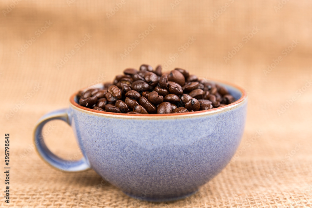 Cup filled with brown roasted coffee beans isolated over burlap very blurry background Cup filled with brown roasted coffee beans isolated over burlap very blurry background