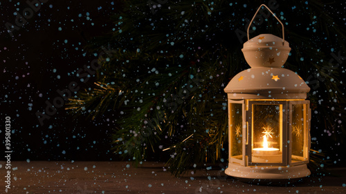 White candle lantern with burning candle. Blurred spruce brunch on the dark background. Falling snow. Retro style picture.