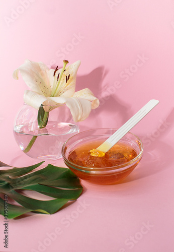 Sugar paste for hair removal in a bowl with lily flower and dracaena leaf in glass vase on pink background, body care