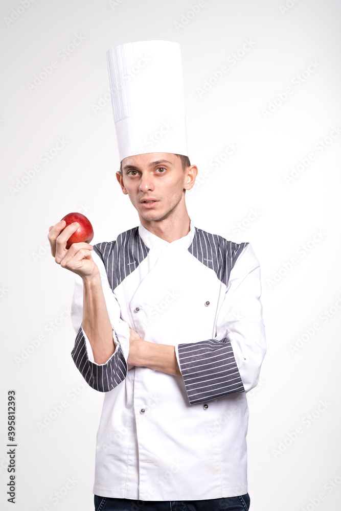 Smiling chef is holding apple on copyspace. Chef showing fruit. Pleased happy young chef posing isolated over white wall background in uniform.