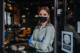 Waitress stands at the open front door of a posh restaurant and waiting for a guests. She wears protective mask as part of security measures against the Coronavirus pandemic. Open for business concept