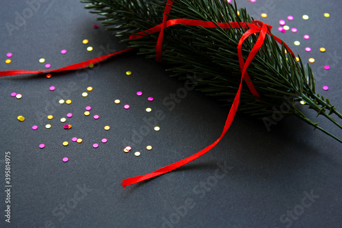 Festive Christmas background with a bunch of spruce twigs, multicolored glitter confetti and bright thin red ribbon. Black backdrop. New Year.