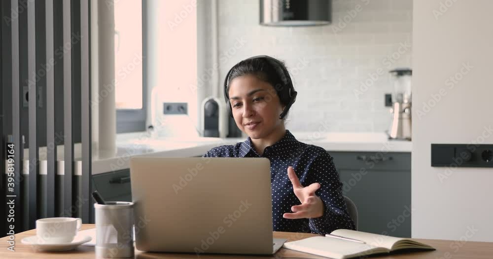 Smiling young pretty indian woman in earphones with mic, enjoying pleasant video call talk with friends on computer, sitting at desk in modern studio kitchen, virtual educational event conference. Smiling young pretty indian woman in earphones with mic, enjoying pleasant video call talk with friends on computer, sitting at desk in modern studio kitchen, virtual educational event conference.