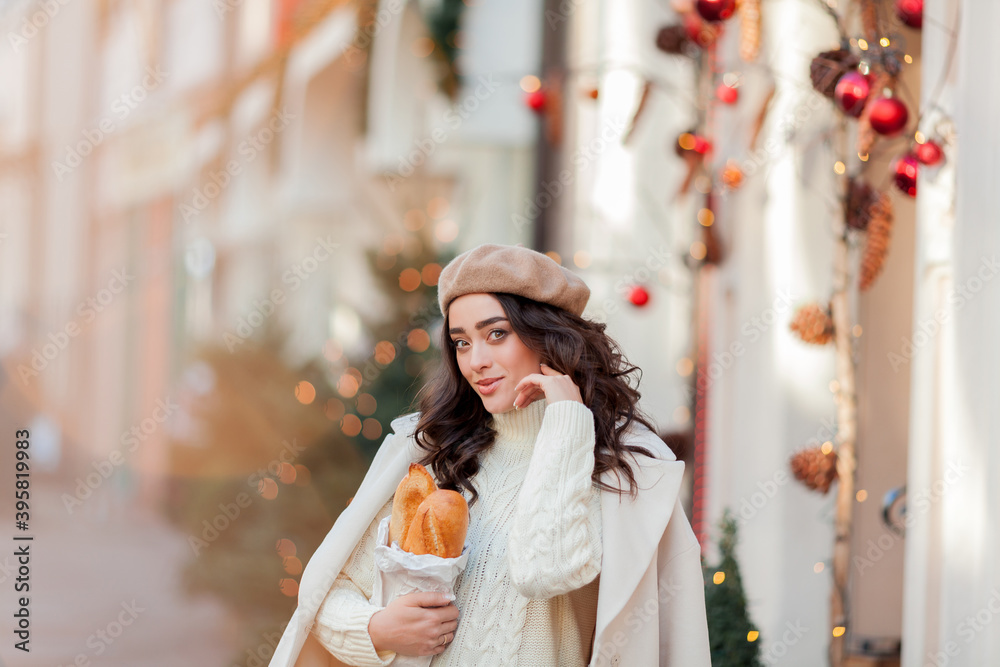 Portrait of a young beautiful woman in a beret in a European city. Young woman holds a paper bag with baguettes. Christmas. Portrait of a young beautiful woman in a beret in a European city. Young woman holds a paper bag with baguettes. Christmas.
