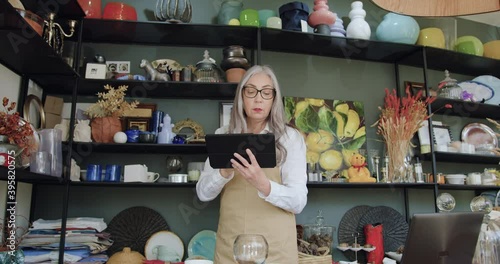 Close up of attractive hardworking concentrated senior woman with long grey hair in glasses which counting goods in gift shop and noticing datas on tablet computer photo