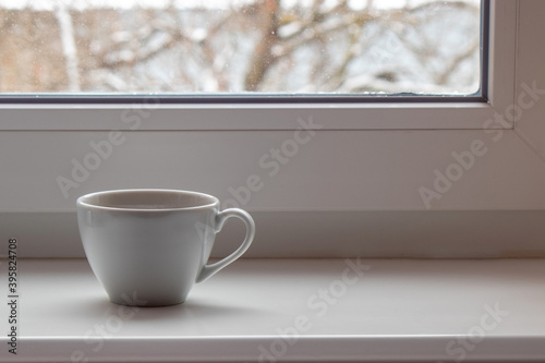 White cup on a white windowsill against the background of a snowy window
