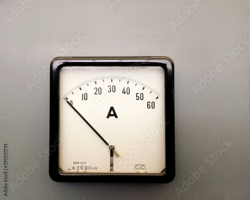 a large square industrial ammeter with an analogue dial with numbers with standard electrical symbols on a white dial on a grey background