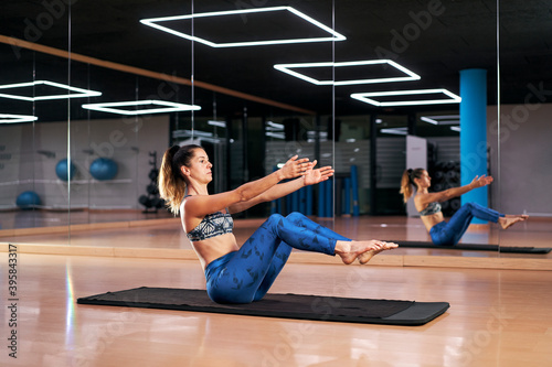 Attractive young woman practicing yoga or pilates in a gym, exercising in blue sportswear, doing boat pose or Agnistambhasana. Full length silhouette.