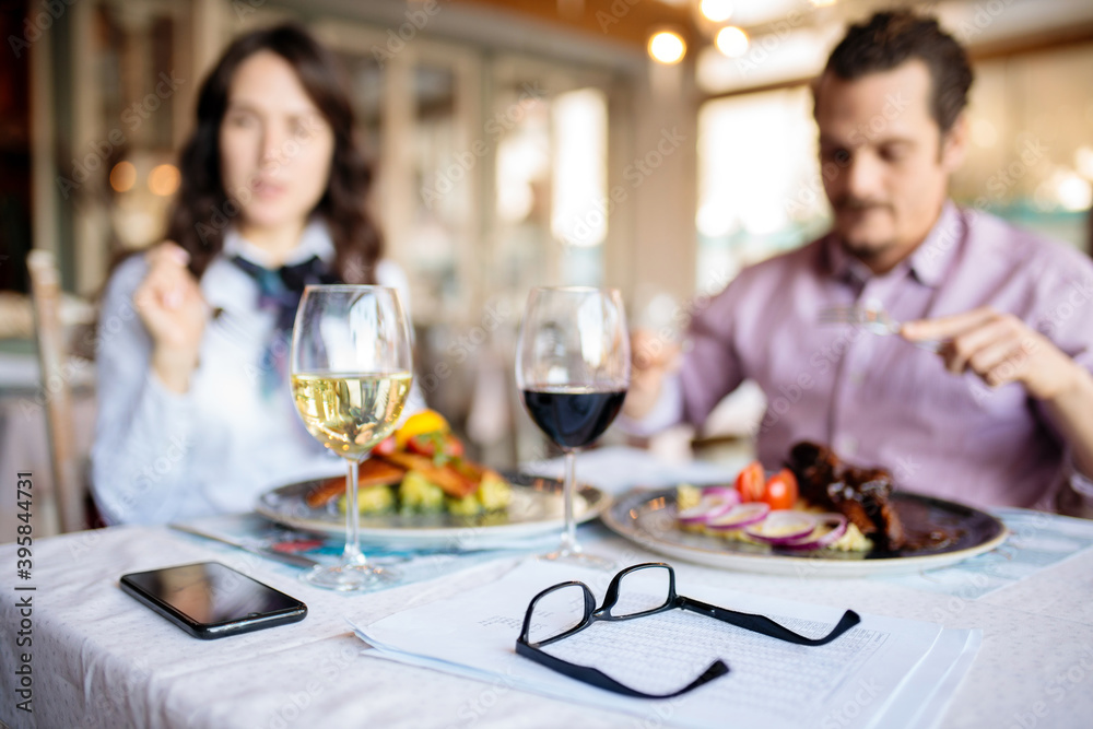 Close-up of eyeglasses resting on report papers on a table in restaurant with unrecognizable male and female business people having lunch in the background