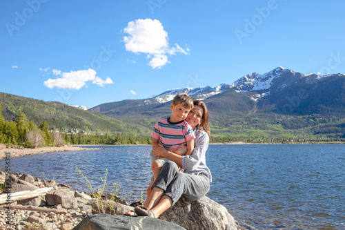 Mother with her little boy sitting on the dock near a beautiful mountain's lake against the background of beautiful mountain landscape. Walk with a child in the fresh air. Happy family concept. 