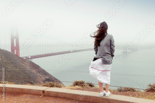 Back view of stylish brunette girl with long hair in jacket and skirt. Woman stands on a hill in foggy windy weather and admires the panorama and view of the bridge. Golden Gate, San Fancisco