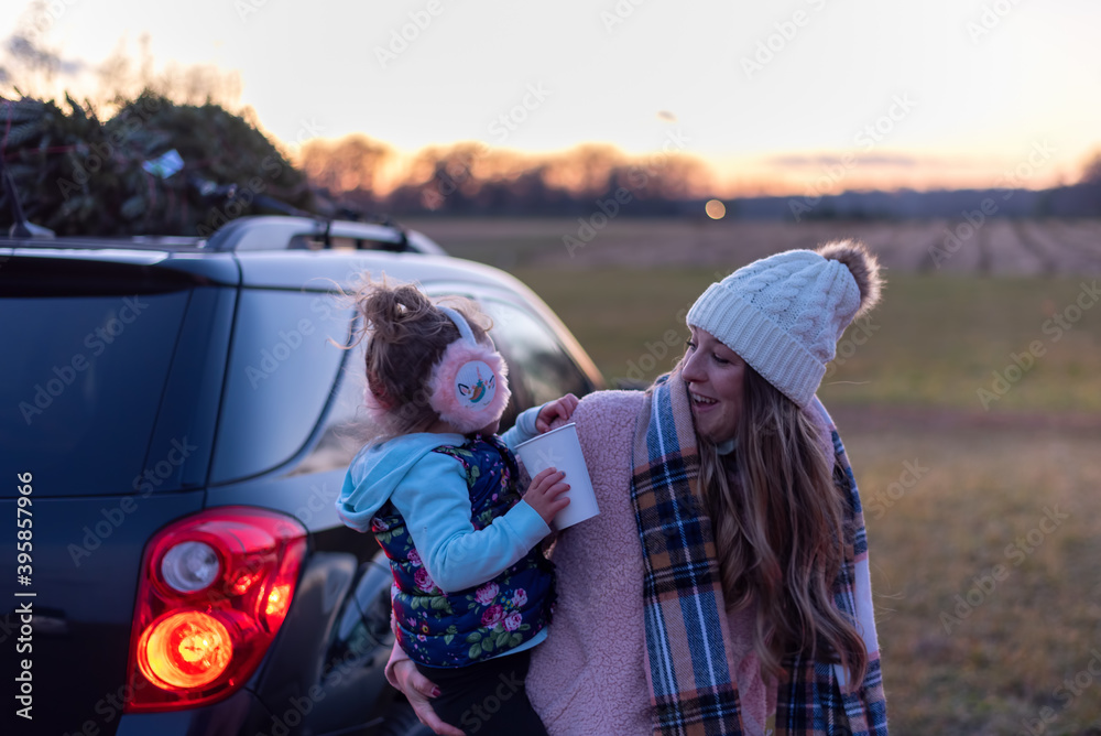 Young mom and little girl at the tree farm with Christmas tree on top of the car