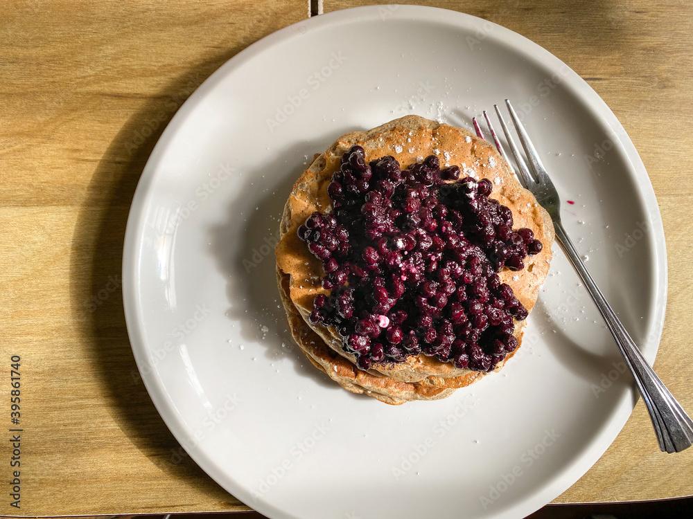 Closeup of a stack of pancakes with a fresh blueberry compote topping on a white plate. From above perspective. Fork on right side. Closeup of a stack of pancakes with a fresh blueberry compote topping on a white plate. From above perspective. Fork on right side.
