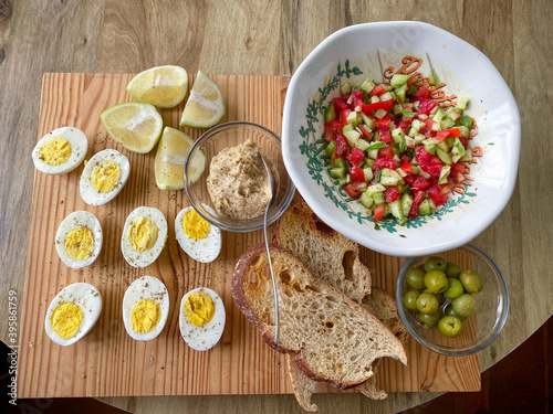 Above view of a Turkish breakfast platter. With halved boiled eggs, sourdough toast, a bowl of diced tomatoes and cucumbers, halved lemons, a small glass bolw of hummus and a bowl of green olives