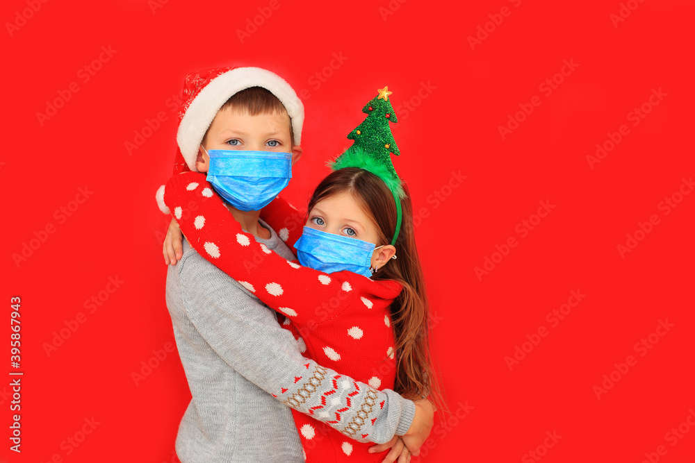 Children in respiratory masks and New Year's outfits hug on a red background during the quarantine period Children in respiratory masks and New Year's outfits hug on a red background during the quarantine period