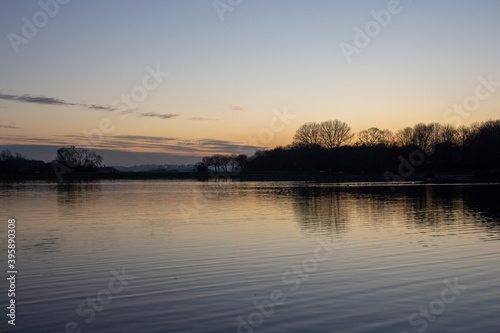 View at sunset with blue sky with some clouds at the horizon reflected on a lake in a cold autumn day, intense orange and dark colors at dusk in Stevenage, Hertfordshire, United Kingdom © BC-Consulting