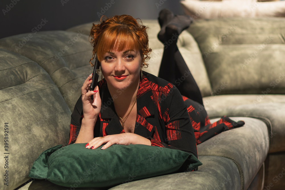 Obraz premium A young red-haired girl lies on a sofa in a dark room, holds a mobile phone to her head and listens attentively to the conversation on the phone
