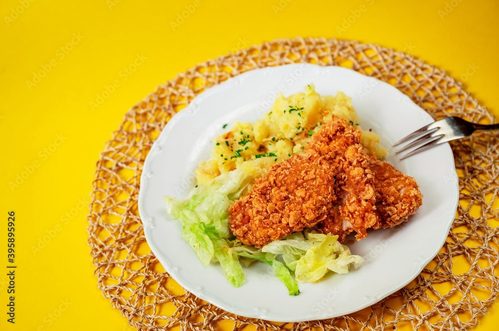 Chicken piece breaded in cornflake, mashed potato and salad in plate, fork on yellow background. Chicken piece breaded in cornflake, mashed potato and salad in plate, fork on yellow background.