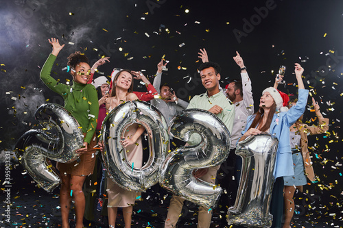 Group of young happy multiracial people holding silver foil balloons in form of numbers 2021 and dancing, confetti falling in the air, celebrating New Years Eve with friends