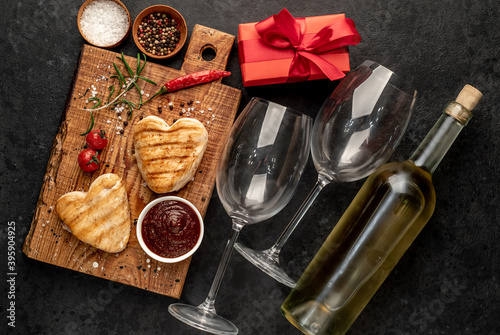 two heart shaped grilled chicken breast steaks with spices, a bottle of white wine with two glasses for Valentine's Day dinner on a stone background