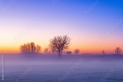 Misty morning in winter with a religious cross and trees in silhouette