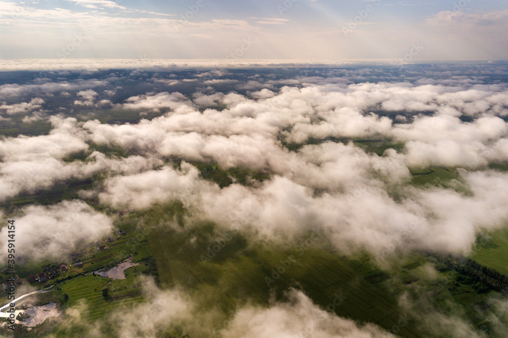 Obraz premium Aerial view of white clouds above a town or village with rows of buildings and curvy streets between green fields in summer. Countryside landscape from above.