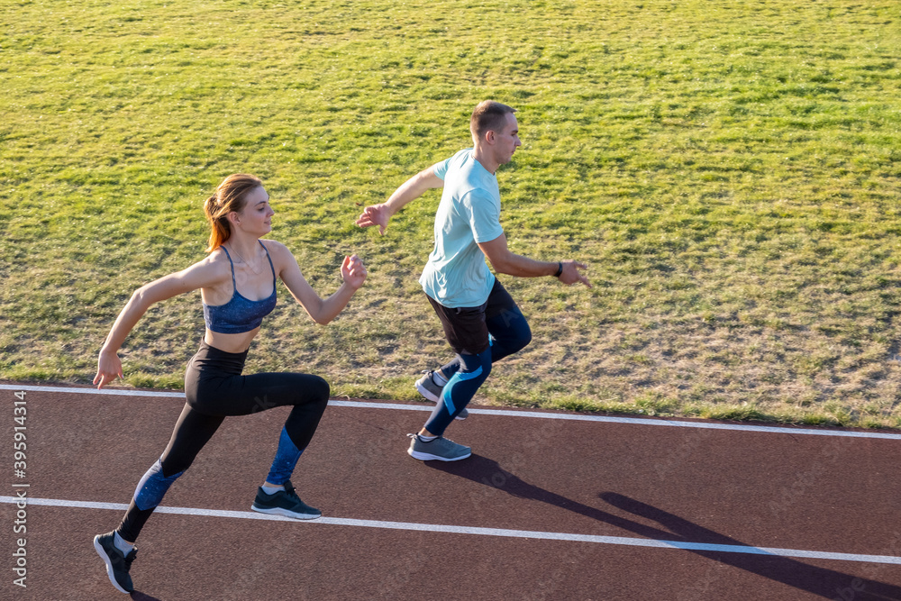 Obraz premium Young couple of fit sportsmen boy and girl running while doing exercise on red tracks of public stadium outdoors.