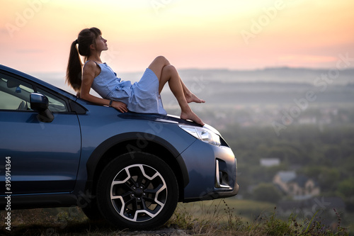Happy young woman driver in blue dress enjoying warm summer evening laying on her car hood. Travelling and vacation concept.