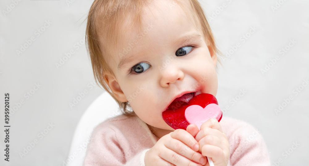 Portrait of a little baby with pink lollipop candy on a white background banner copy space. A child licks a heart-shaped candy. Harm to teeth. Portrait of a little baby with pink lollipop candy on a white background banner copy space. A child licks a heart-shaped candy. Harm to teeth.