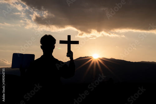 Silhouette of human kneeling down praying and holding christian cross for worshipping God at sunset background. Christian, Christianity, Religion copy space background.