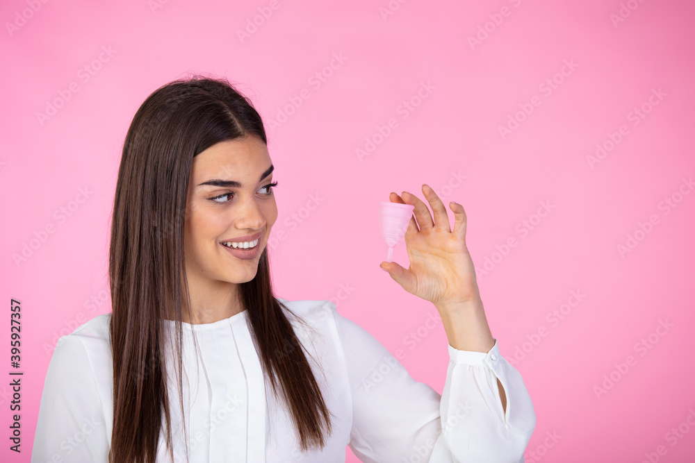 Attractive woman holding menstrual cup standing against pink background, squeesing her shoulders and spreading hand aside, looks smiling at camera. Female period concept. Attractive woman holding menstrual cup standing against pink background, squeesing her shoulders and spreading hand aside, looks smiling at camera. Female period concept.