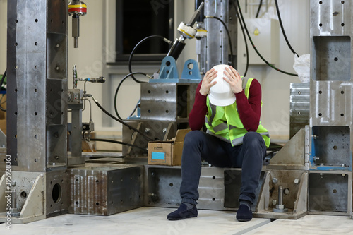 Portrait of a male factory manager in a white hard hat and red sweater holding his head. Solving factory problems. Worker lost the job. Unemployment .